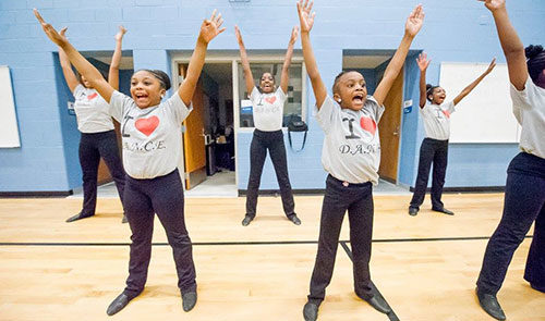 Several children wearing "I heart dance" t shirts smile widely and raise their arms in the air in the form of a "V"