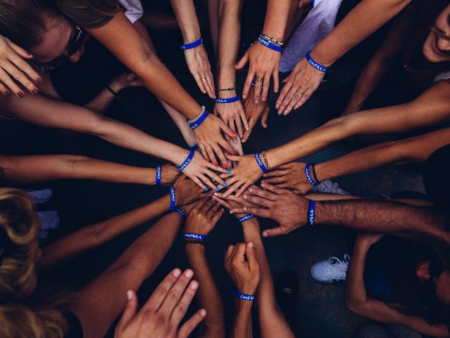 A circle of many people wearing “Emfasis” bracelets outstretch their hands to meet in the center of the circle.