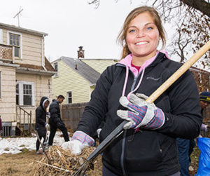 A girl smiles holding a rake full of leaves in her gloved hands.