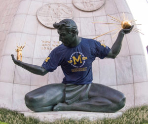 Statue of a man sitting cross-legged holding a sun in his right hand wearing a blue t-shirt with a block M on it.