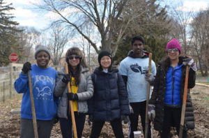 Five volunteers stand outdoors, dressed in hats and coats, holding rakes, and smiling.