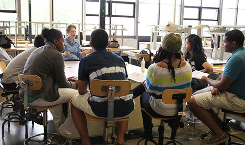 Students around a table listening to instructor.