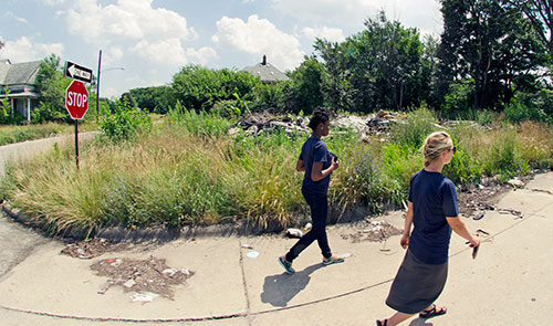 Two people walk down a Detroit sidewalk next to a green patch of grass on a sunny day.